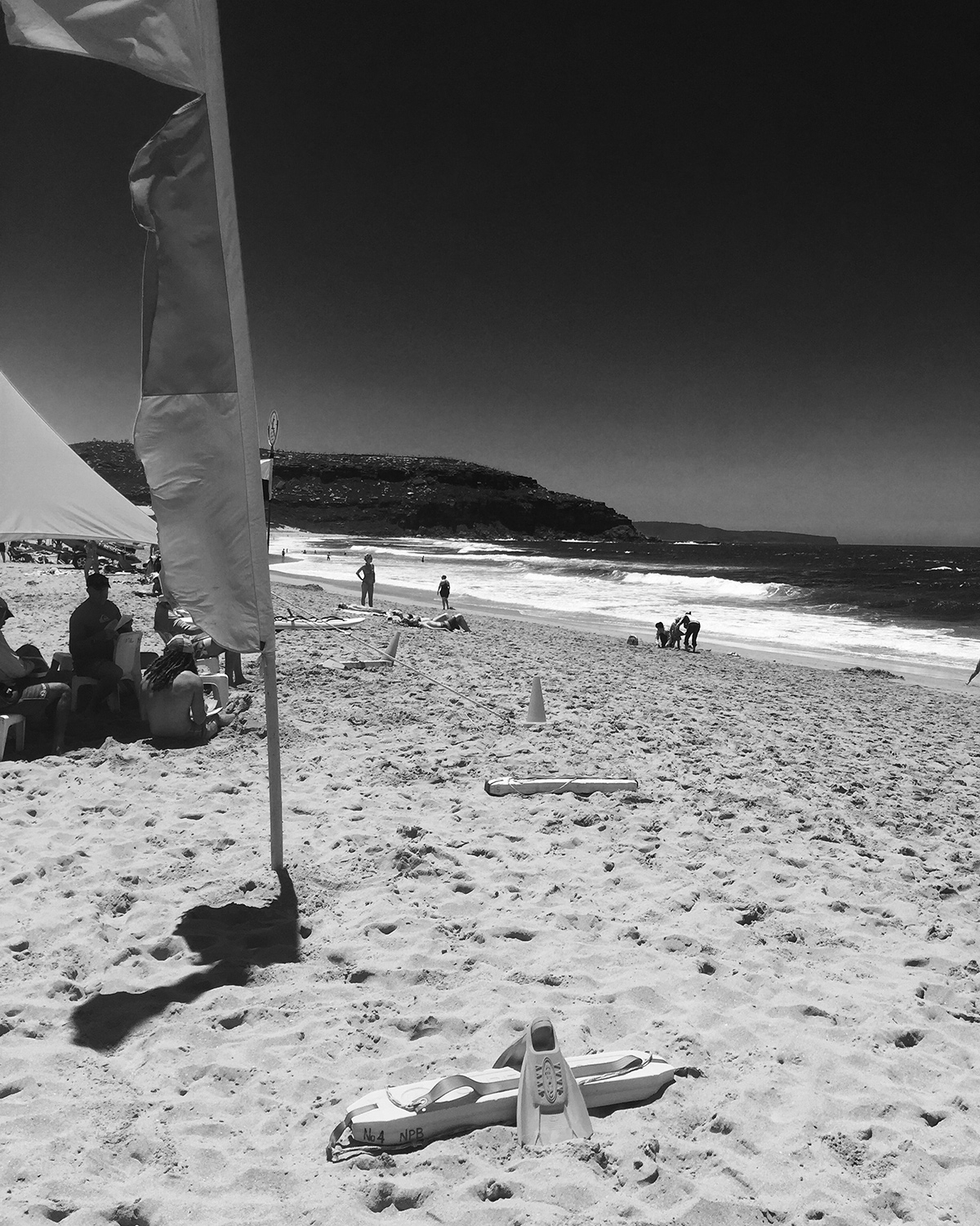A black and white image of the Surf Lifesaving flags and groups of people sitting along the beach 