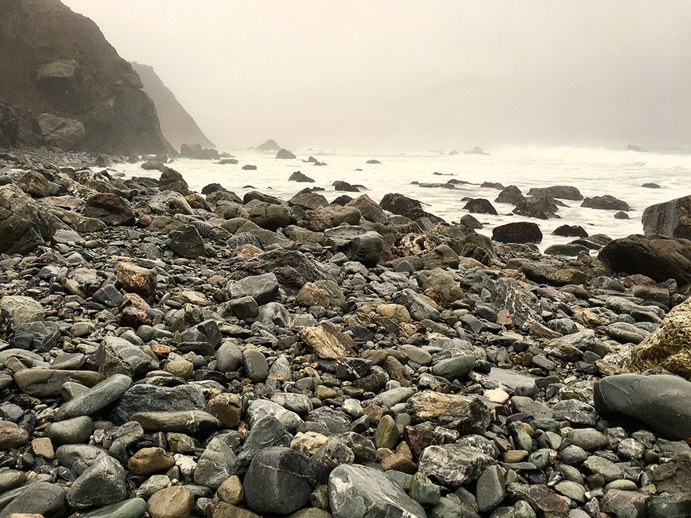 A rocky shore and whitewashed water sits underneath a hazy cloud 