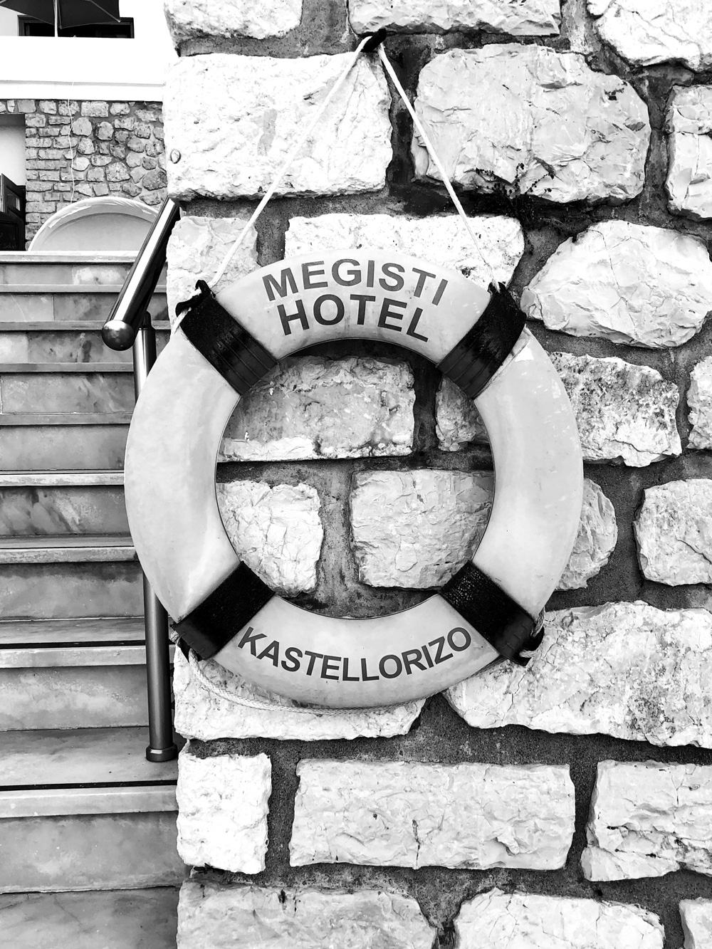 A black and white image of a round buoy hanging off a stone wall at Megisti Hotel that reads “Megisti Hotel Kastellorizo” 