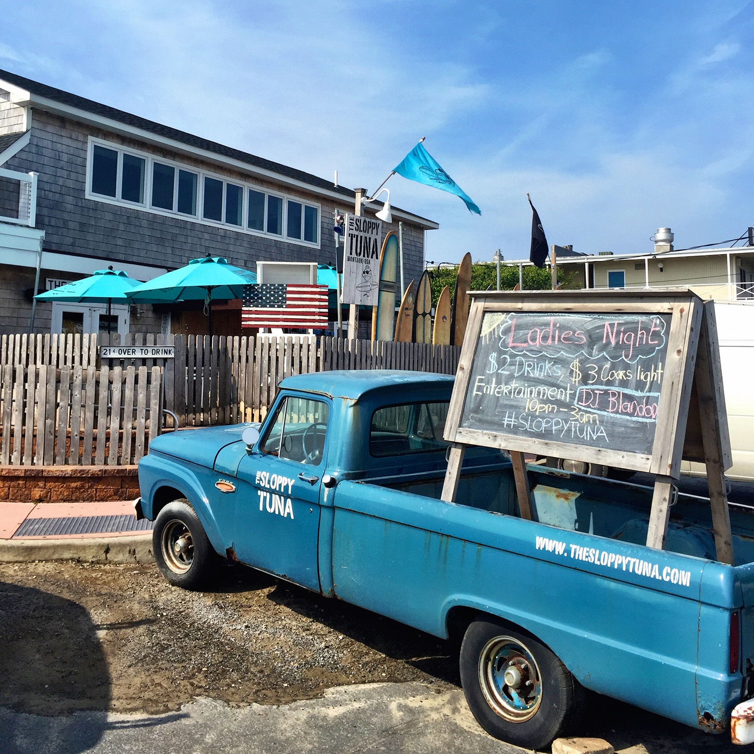 A blue pickup truck parked on the sidewalk in front of a shingle house has a large blackboard sign in the tray advertising a Ladies Night 