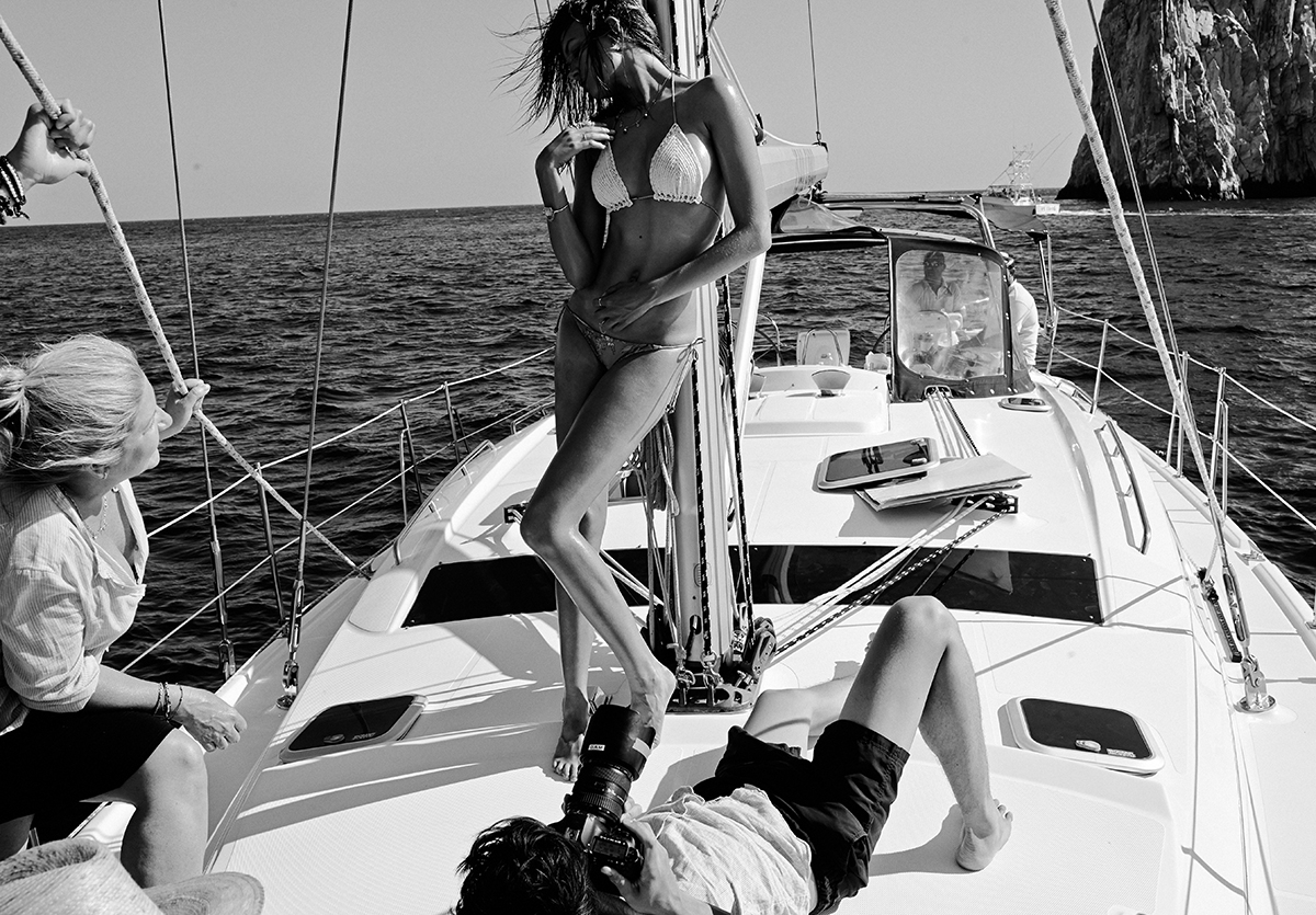 A black and white behind the scenes image of our model standing against a pole on a yacht with the photographer lying at her feet and another crew member watching on 