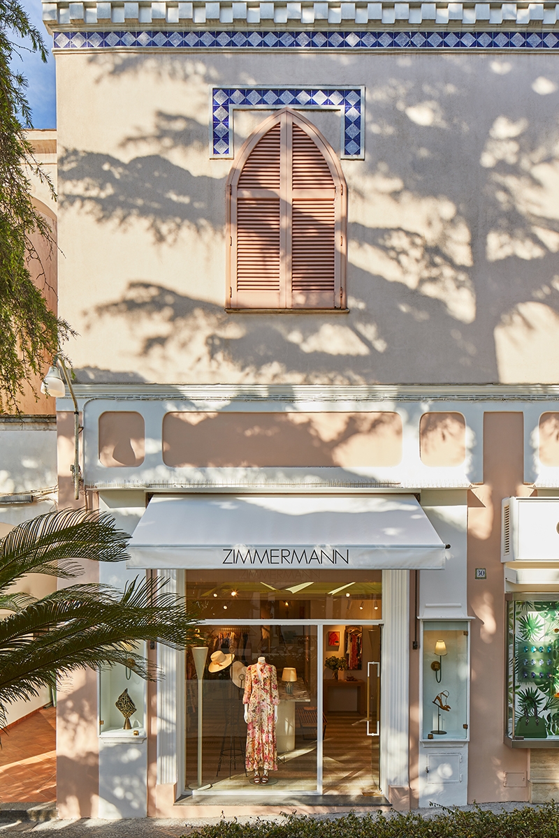 The exterior of our Capri boutique displays a muted apricot building with white finishes and blue tile detailing. A single mannequin stands in the window and two casings of accessories feature on both sides of the entrance. 
