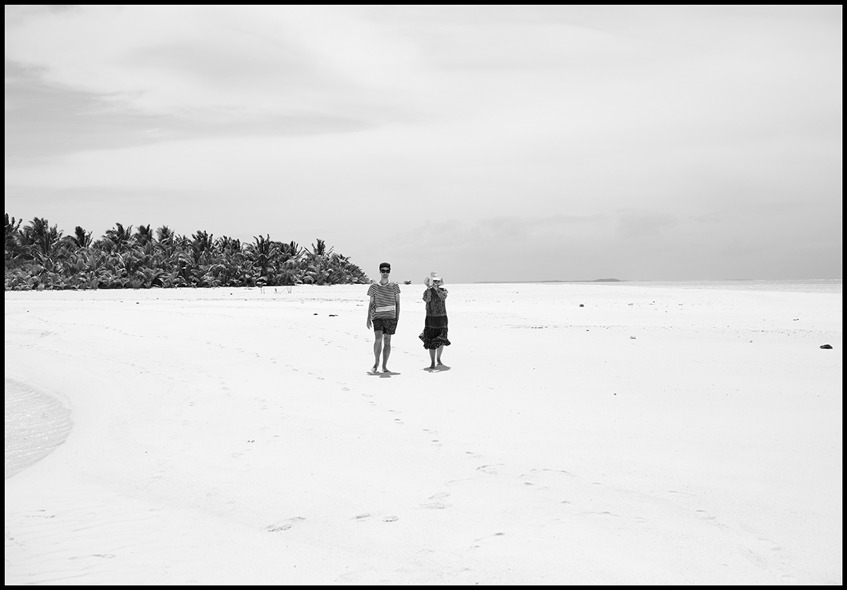 A black and white image of two people standing along expansive white sands, with tropical palms clustered in the background  