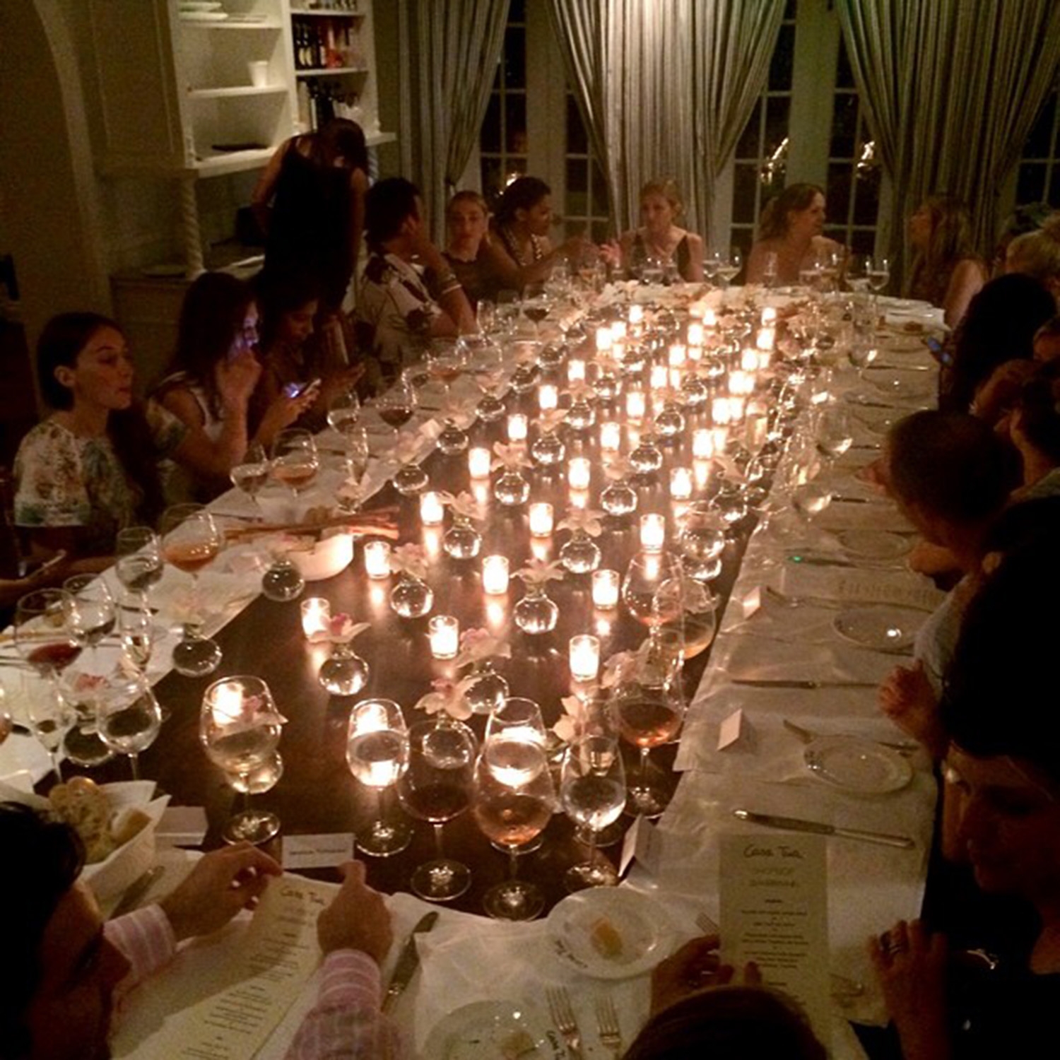 A group sits around a large rectangle dinner table that is lined with plates, wine glasses and candles 