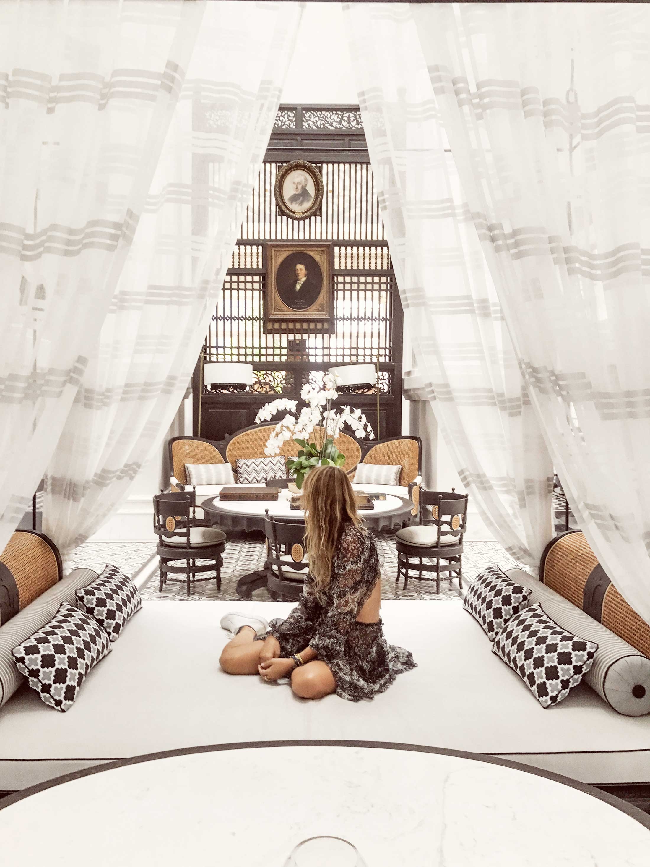 Romy sitting on a large canopy bed with luxurious white curtains and patterned pillows
