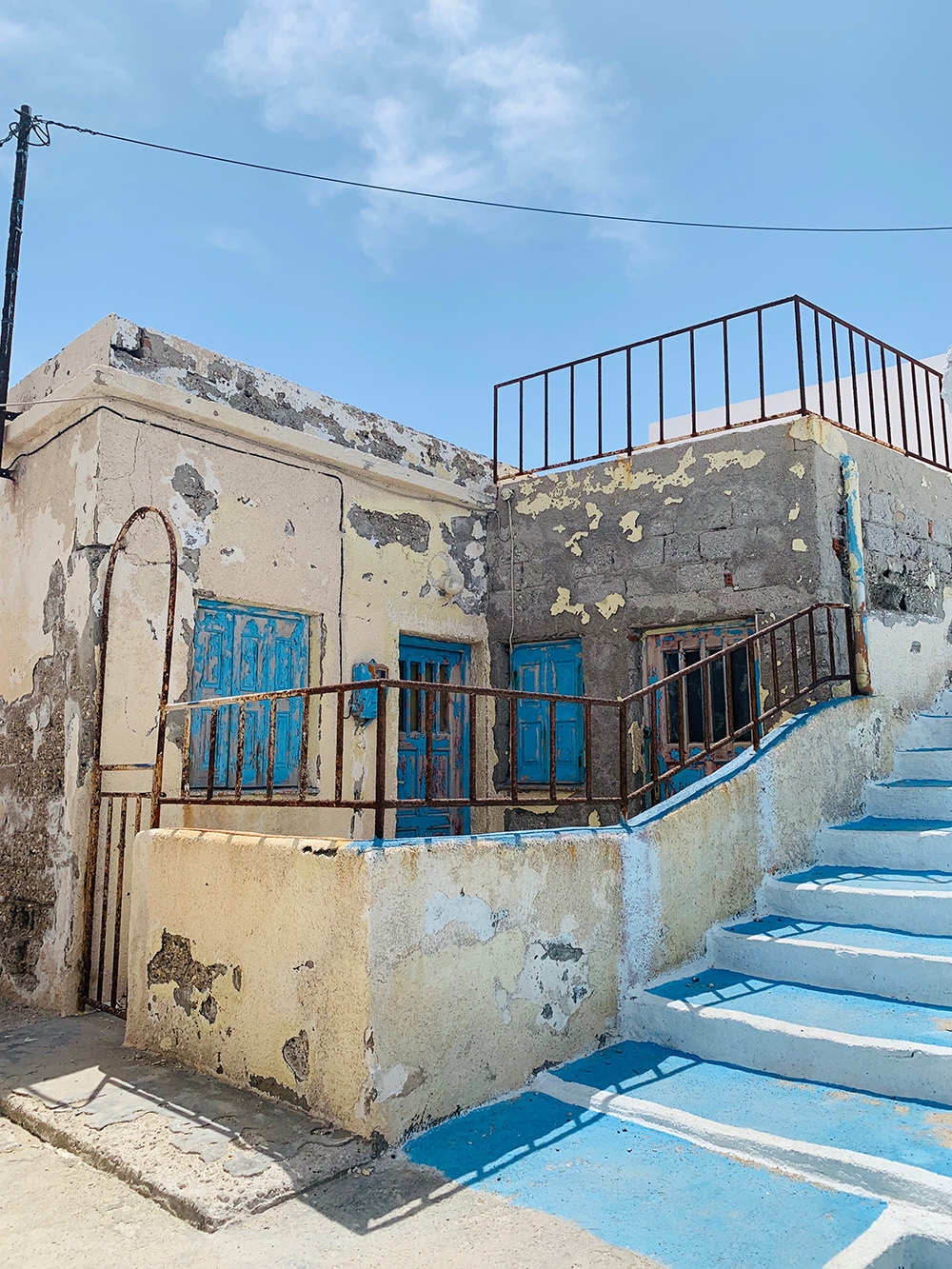 An old Greek building with peeling cream and blue paint on the walls, doors and railings. A blue staircase ascends to the second level. 
