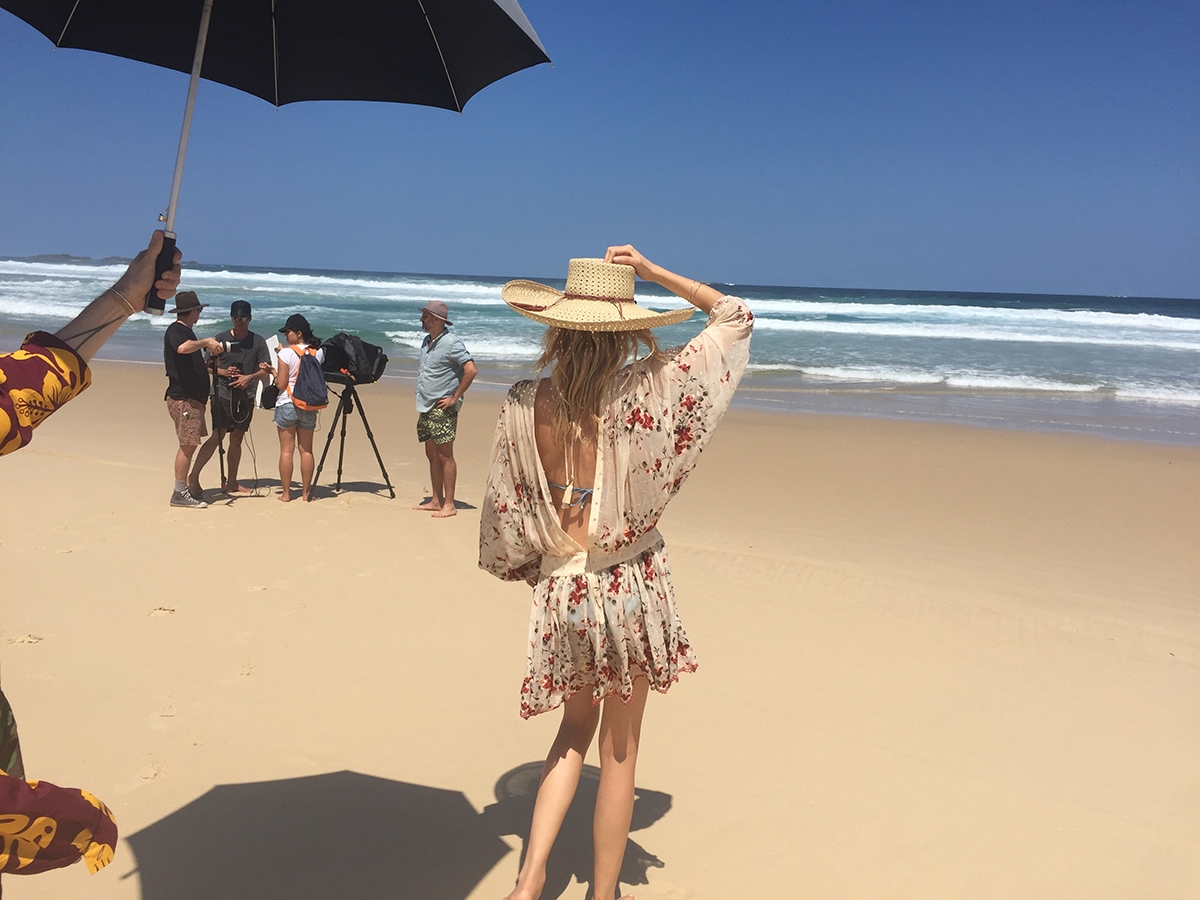 A behind the scenes shot of Lily Donaldson waiting under an umbrella while the crew chat on the beach 
